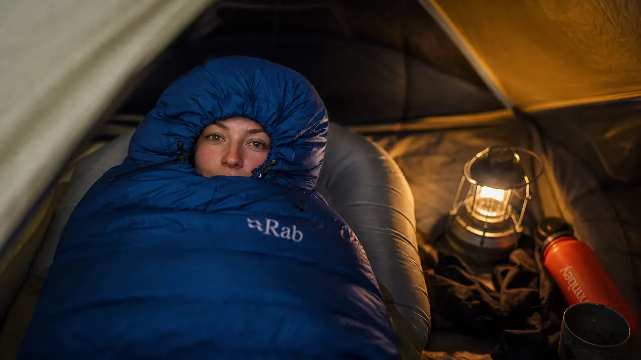 Camper sleeping soundly in a mummy bag under a star-filled sky with tent walls glowing from a lantern inside