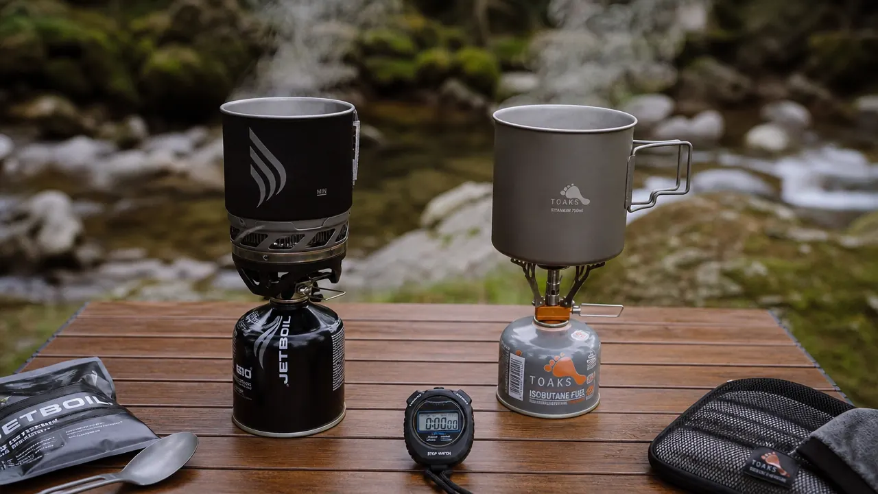 Backpacker cooking breakfast at camp using a small canister stove, mist rising from a mountain lake in the background